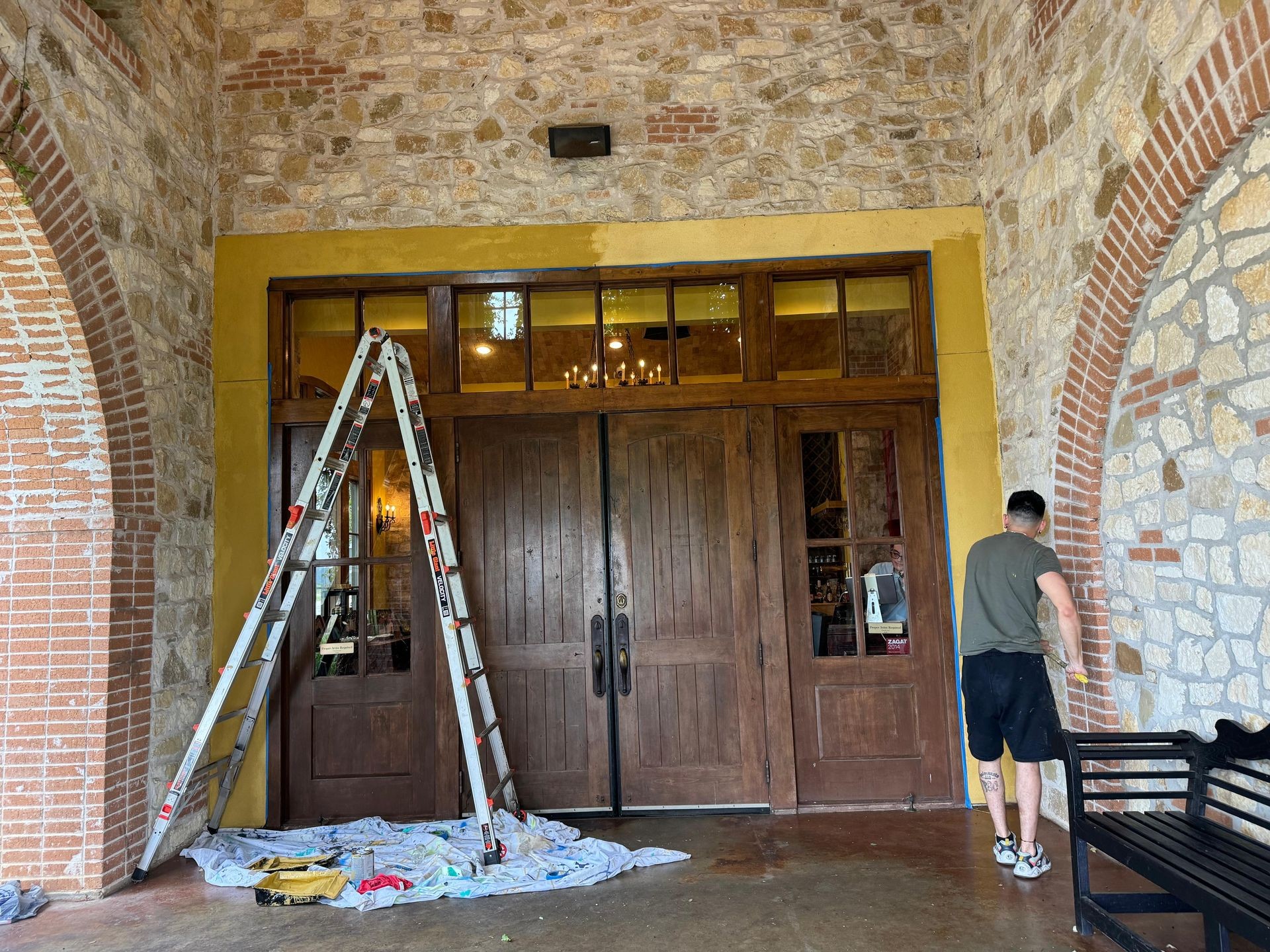 Person painting a stone wall near a large wooden door with ladder and painting supplies on the ground.