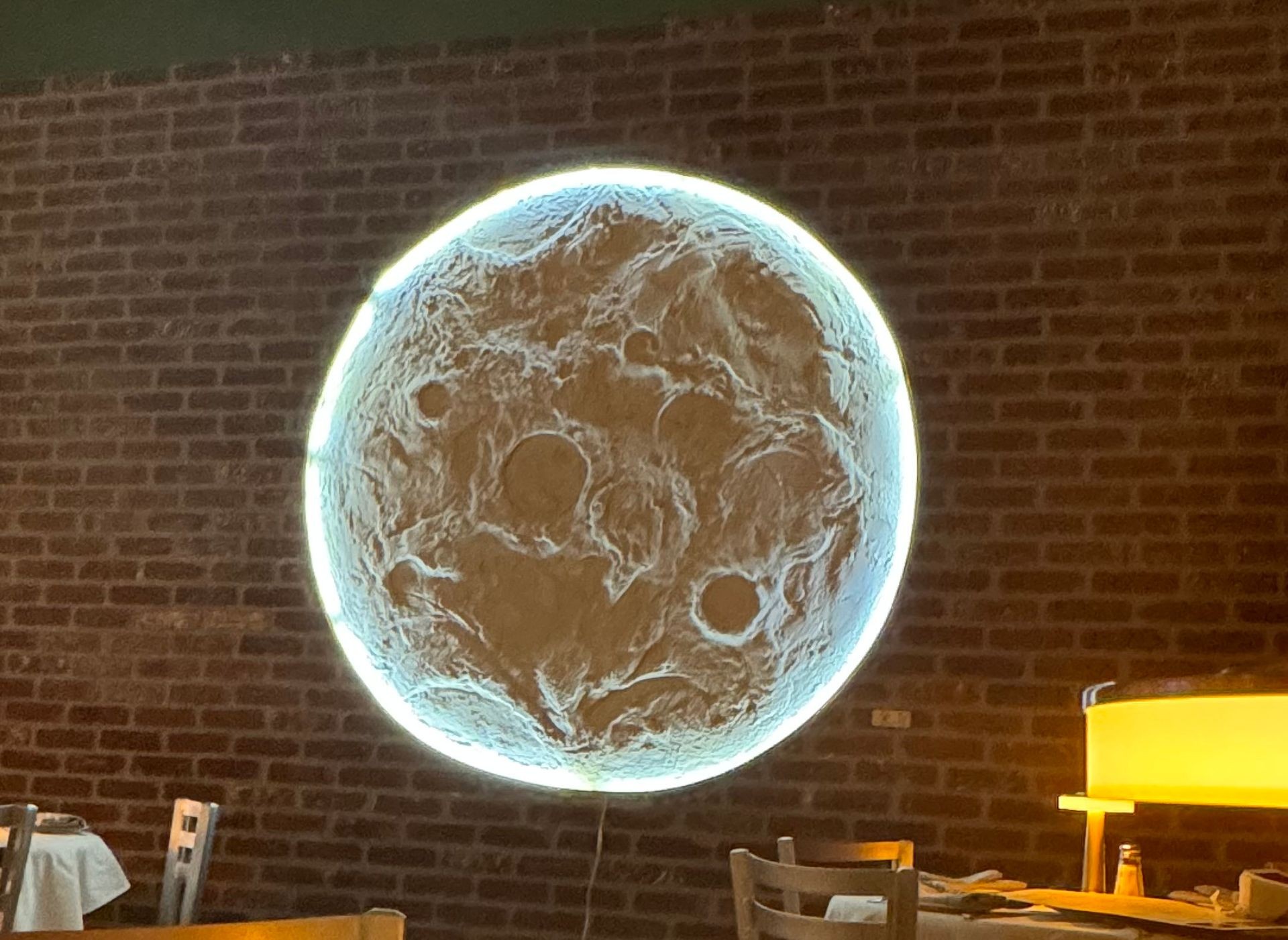 Restaurant interior with a circular, illuminated moon-shaped wall decor against a brick wall.